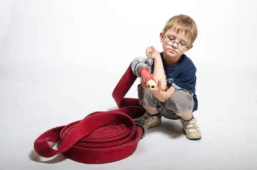 A young boy wearing glasses looking closely at a heavy red fire hose to learn how to stay safe and not get hurt.