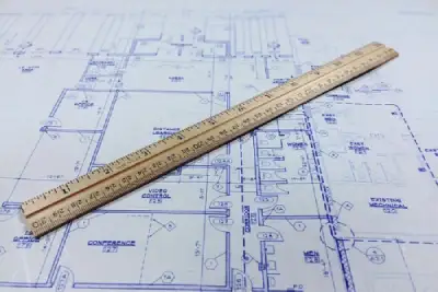 A wooden ruler placed on top of a blue house floor plan or blueprint for precise measurement.