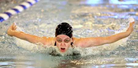 A female swimmer in a black cap and goggles performing a powerful butterfly stroke in a swimming pool.