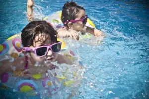 Two young girls wearing sunglasses and using colorful inflatable rings while swimming in a bright blue pool.