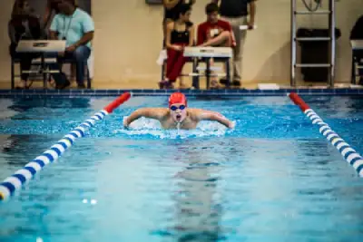 A male swimmer with a red cap and goggles racing butterfly stroke down a lane during a swimming meet.